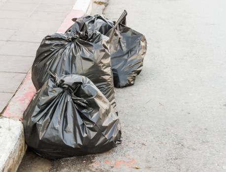 Three large black plastic rubbish bags, filled and tied at the top, are placed on a paved surface near a street curb. The bags are slightly crinkled and appear to contain garden or household waste. One bag leans against the curb while the other two are positioned close together on the asphalt, with a small stick partially visible among them. The surface below is smooth and grey, with a red painted line marking the edge of the pavement. The background shows the edge of the pavement and part of the road, indicating an outdoor urban setting suitable for local rubbish removal services, such as those offered by Garden Clearance Enfield in the Enfield area.