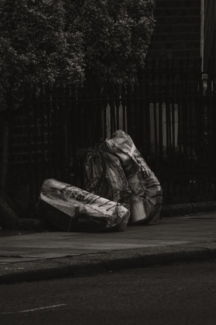Two large black garbage bags filled with rubbish, leaning against a black metal fence along a paved sidewalk in a residential area. The bags appear tightly packed and slightly crumpled, with some visi