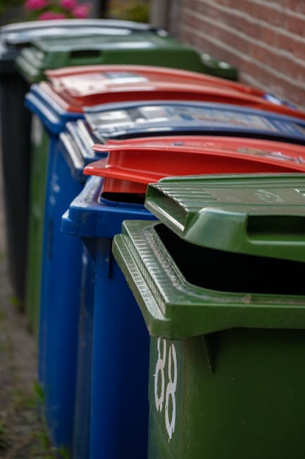 Two large black garbage bags filled with rubbish, leaning against a black metal fence along a paved sidewalk in a residential area. The bags appear tightly packed and slightly crumpled, with some visi