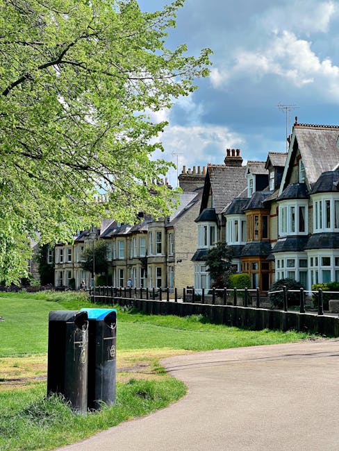 A residential street scene featuring a row of traditional terraced houses with bay windows and slate roofs situated behind a well-maintained green lawn. In the foreground, there are two black public r
