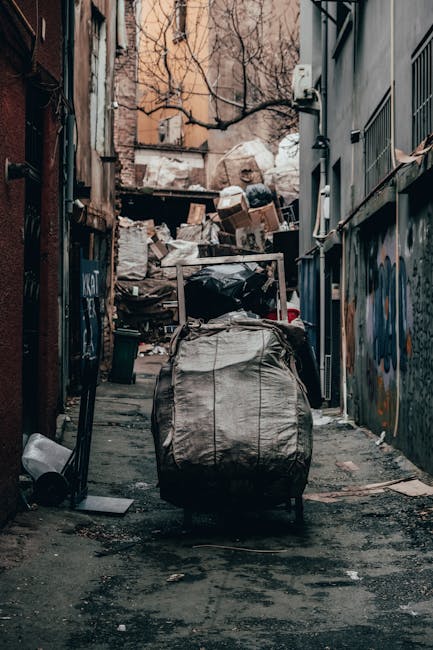 Two large black plastic bags filled with waste materials are placed on the curbside pavement next to a metal fence and dense greenery. The bags appear to contain household or garden rubbish, with one 